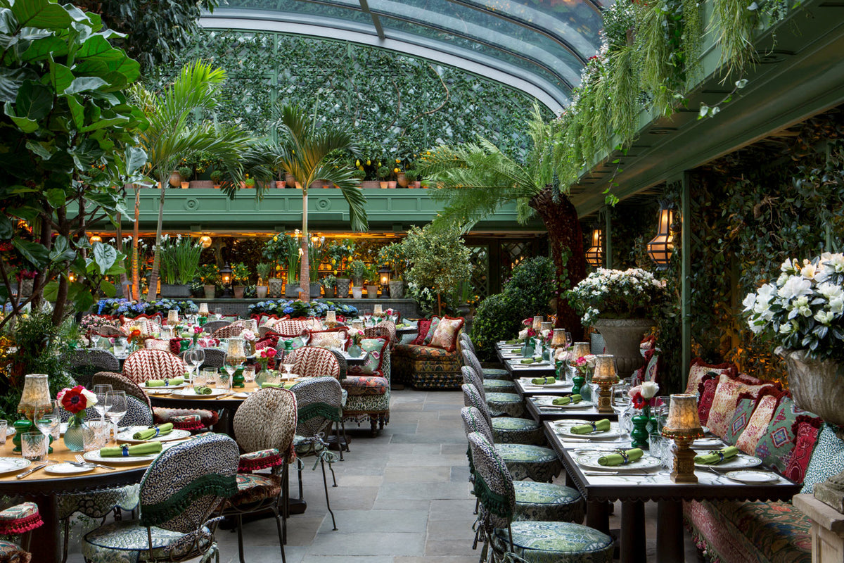 A hospitality project furnished by Decca. A modern restaurant with chairs and benches upholstered in green and red floral fabric. The walls are covered in vines and there are palm trees spread all over the establishment.