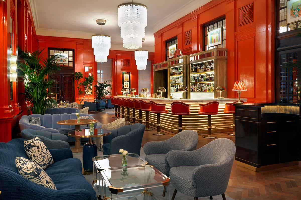 A modern hospitality interior furnished by Decca. It features red wooden walls, a gold-toned bar with red bar chairs and blue sofas and armchairs around glass tabletop coffee tables.