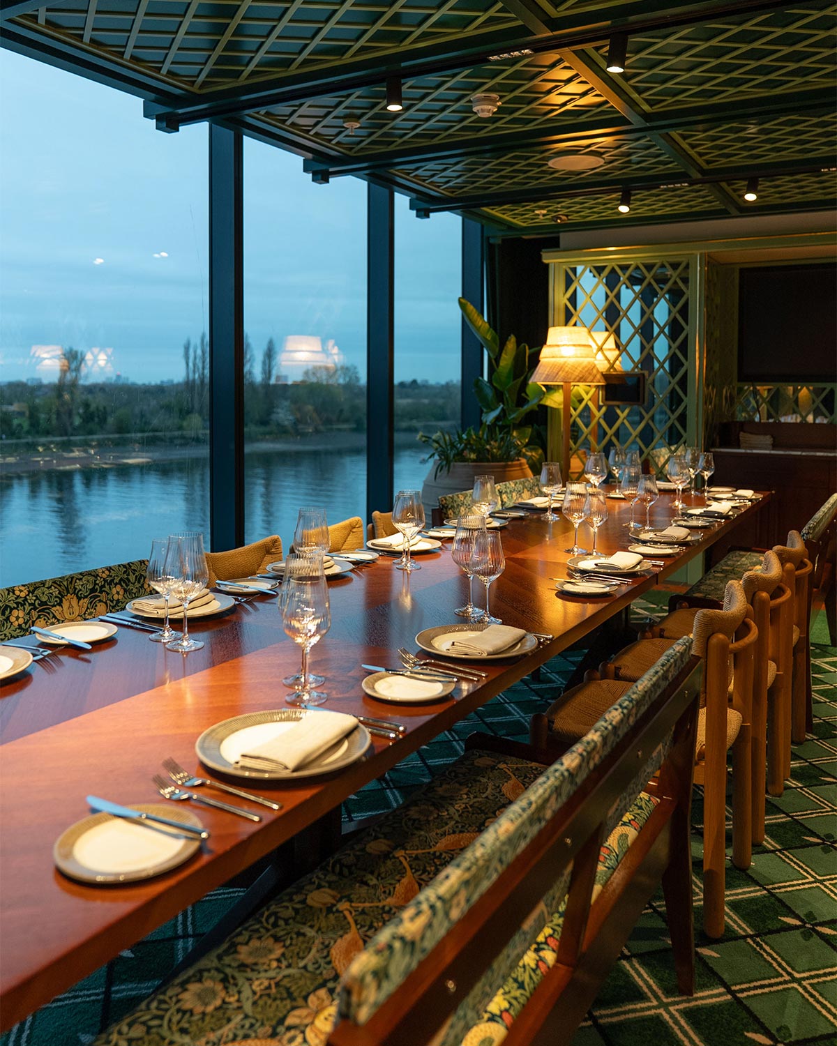 The Members Club at Fulham Pier, furnished by Decca. A long wooden dining table fully set with plates, wine glasses, and cutlery, positioned in a luxurious riverside restaurant with large windows overlooking the water at dusk.