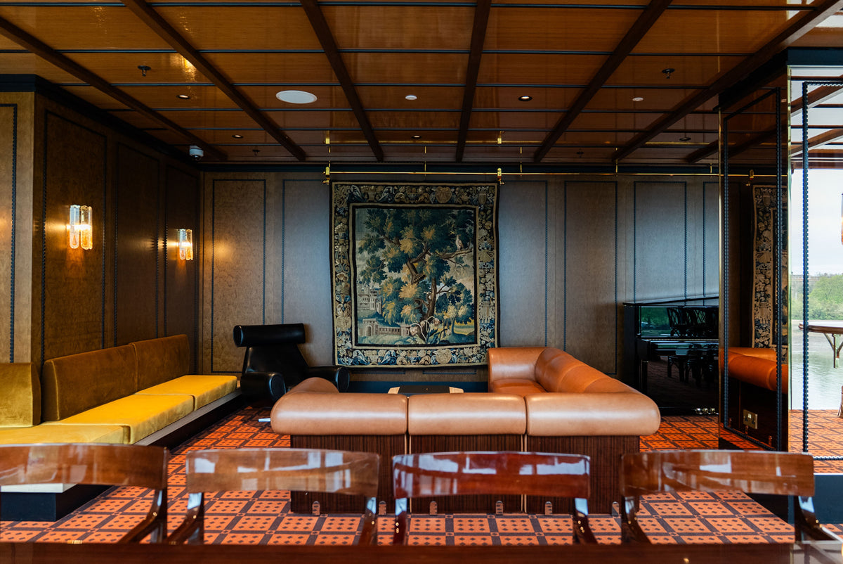 The Members Club at Fulham pier, designed by Stuart Forbes Associates, furniture manufactured by Decca. Modern interior with a large brown leather upholstered sofa, a black leather armchair, and against the wall, a light brown upholstered bench. In the foreground is a large wooden table with chairs.