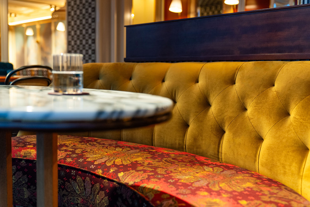 A close-up of a luxury tufted velvet booth in mustard yellow, paired with a red and gold patterned upholstered bench and a round marble-top table in a warmly lit indoor setting.