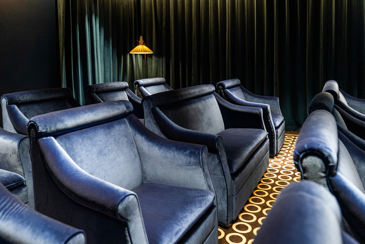 A row of plush dark blue velvet armchairs arranged in rows inside a dimly lit room with dark green curtains and a patterned gold and black carpet.