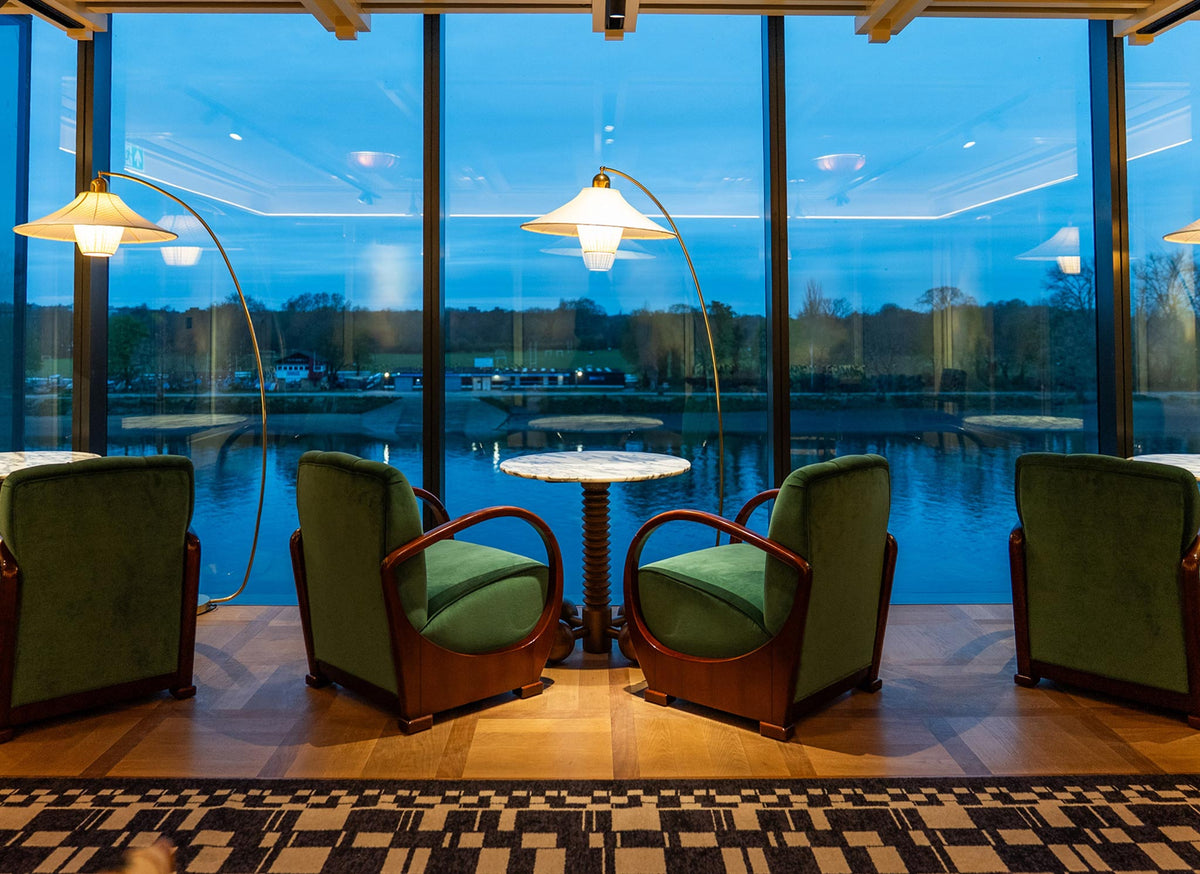 The Members Club at Fulham Pier - A hospitality project manufactured by Decca, designed by Stuart Forbes associates. A modern lounge area with four green upholstered armchairs arranged around a small round table, set in front of large floor-to-ceiling windows overlooking a calm lake at dusk.