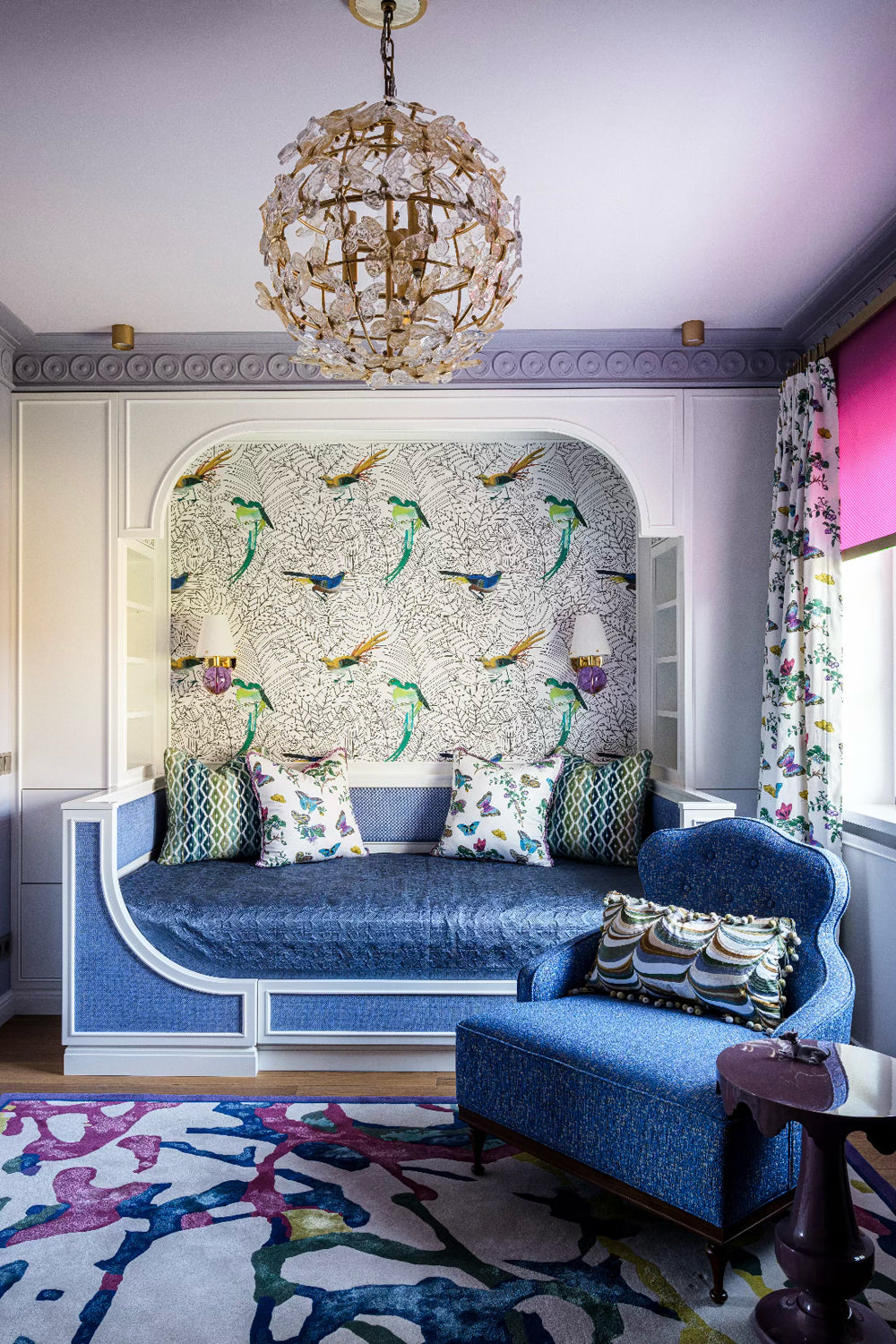 Bedroom with a blue fabric upholstered daybed with matching armchair and round purple side table, colourful pillows with patterns and butterflies in blue and green, and a patterned wallpaper depicting birds.