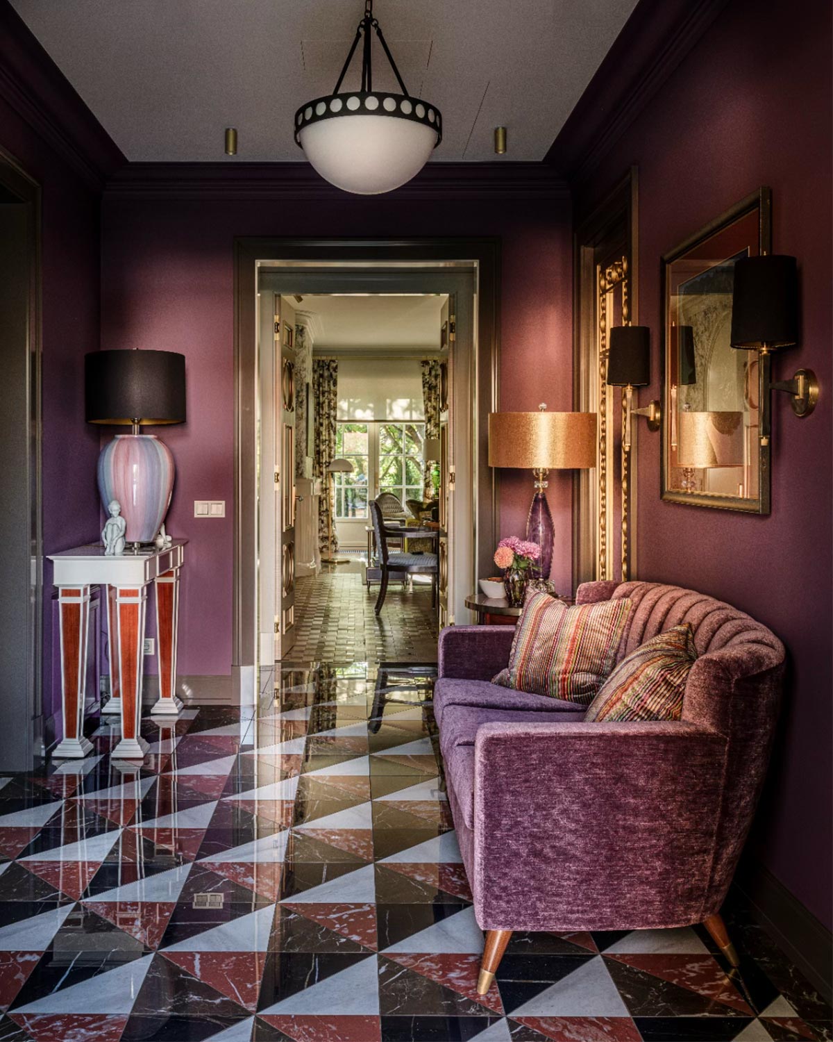 Project Rosinka, a residential project furnished by Decca. A luxurious interior hallway with deep purple walls, a matching purple velvet sofa with patterned cushions, a black chandelier, and a polished marble floor with a geometric black, white, and reddish pattern.