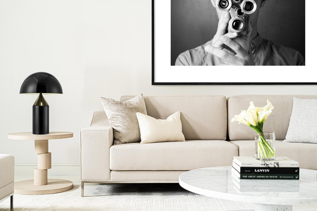 The Domicile new Edition collection, designed by Michael Vanderbyl, manufactured by decca. Pictured is a modern interior with a white upholstered sofa, a light brown wooden side table with a black table lamp, and in the foreground, a white marble table with books and a vase with flowers on top.