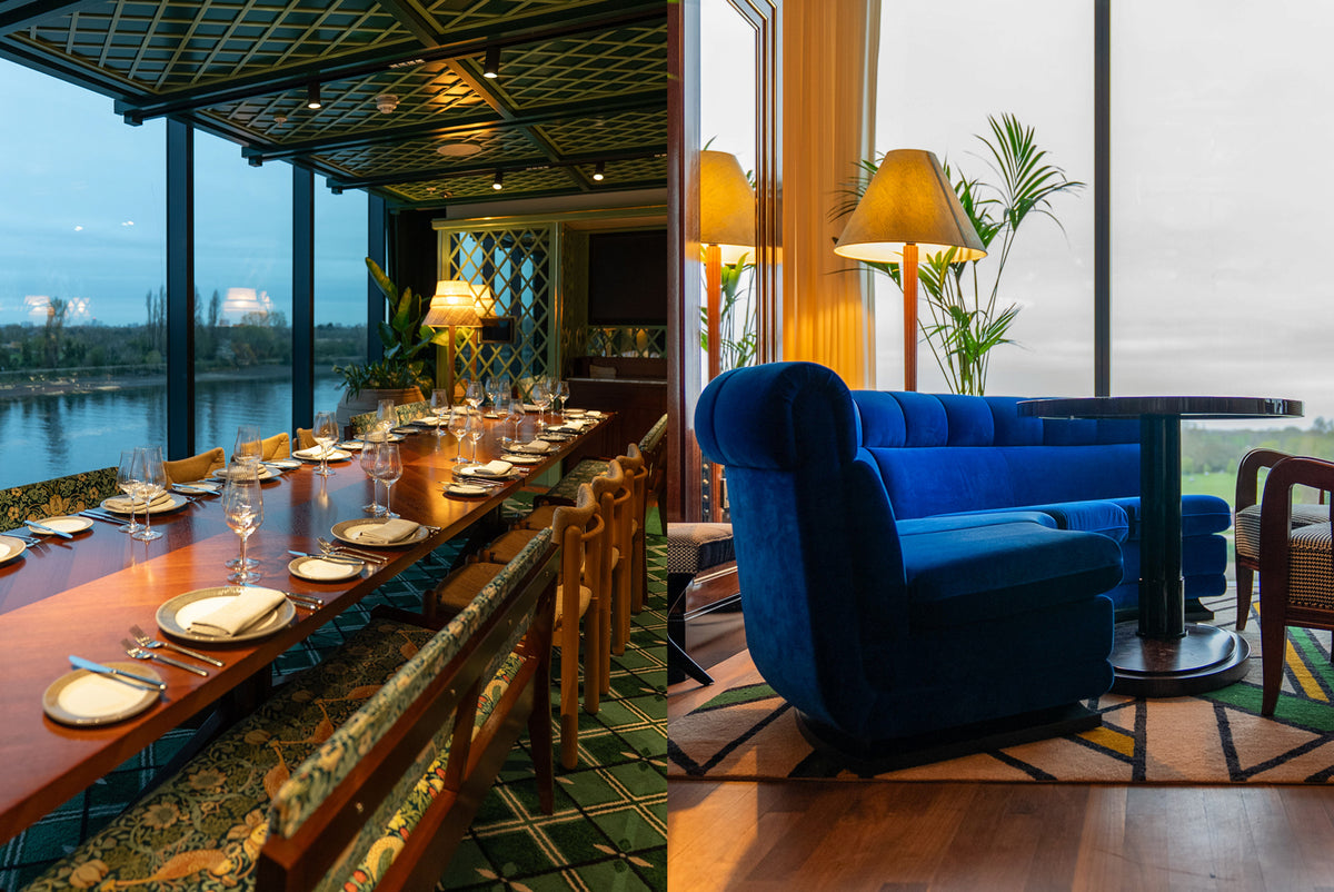  Long dining table with plates and cutlery with benches upholstered in a green floral pattern and chairs on either side, Blue sofa in a room with large windows.