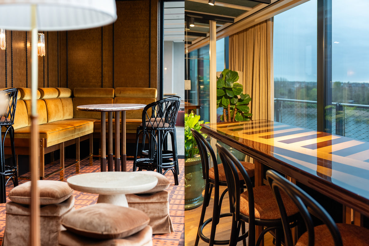 A modern indoor bar seating area with a long wooden bar, black bar stools, mustard yellow upholstered seating, small round tables, and large windows overlooking The Thames