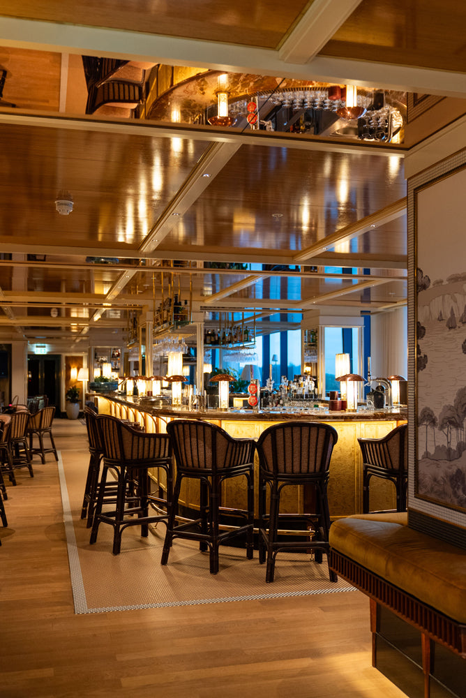 A warm, upscale bar and lounge interior with wooden ceiling panels, bar stools, a lit bar counter, and a yellow upholstered bench in the foreground.