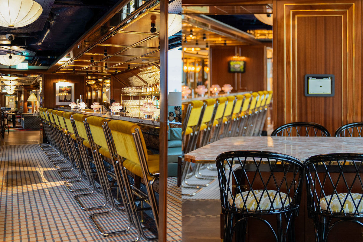 A split-view interior scene of a bar with warm wood paneling, yellow upholstered bar stools, black dining chairs with cream cushions, and a marble tabletop.