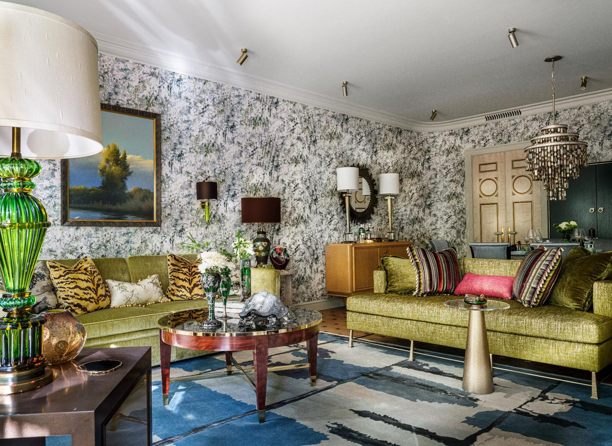 Project rosinka - A residential project designed by Boris Dmitriev with furniture manufactured by Decca. A styled living room featuring two olive green upholstered sofas with patterned and striped pillows, a blue and white abstract area rug, floral wallpaper, and a large green glass table lamp in the foreground.